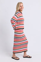 A woman modeling a long-sleeve knit maxi dress with horizontal wavy stripes in red, orange, pink, black, and light blue, shown from a side-back angle against a light gray studio background.