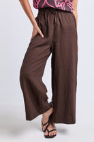 A person wearing loose, wide-leg brown pants with a drawstring waist and side pockets, paired with black strappy sandals against a light gray studio background.