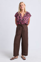 A woman modeling a short-sleeve blouse with a pink and black abstract pattern paired with brown wide-leg drawstring pants and black flip-flops, photographed against a light gray studio background.