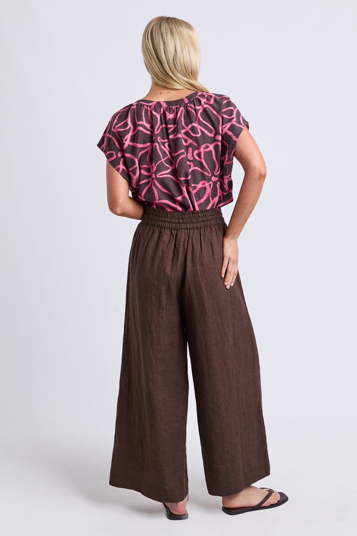 A woman shown from the back wearing a short-sleeve top with a pink and black abstract pattern and high-waisted, wide-leg brown pants, standing against a light gray studio background.
