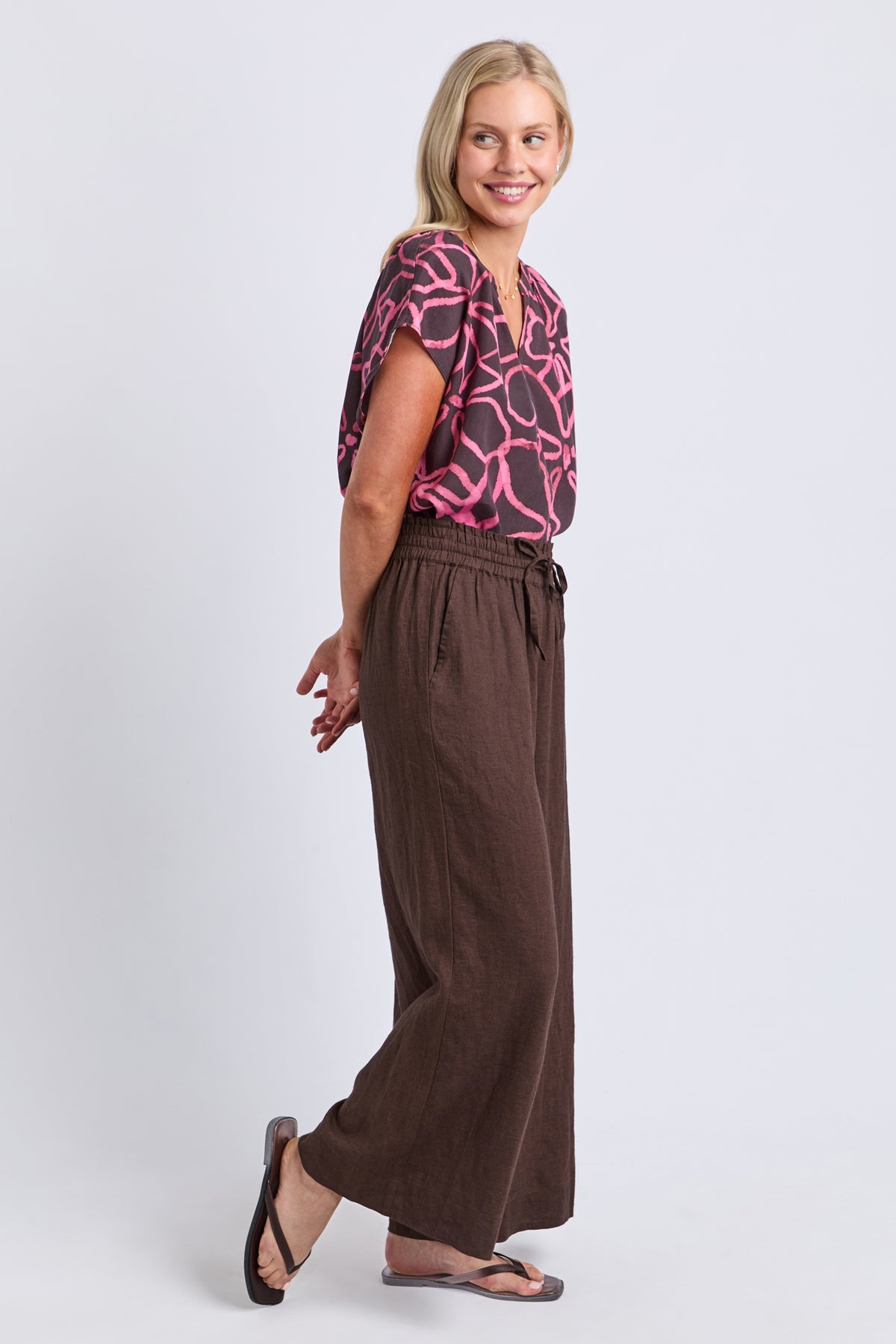 A woman modeling a short-sleeve top with a pink and black abstract pattern and high-waisted, wide-leg brown pants, standing against a light gray studio background.