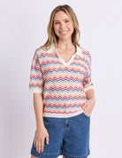 A woman modeling a short-sleeve knit polo-style top with a multicolor chevron pattern in pink, blue, orange, and cream, paired with denim shorts against a light gray studio background.