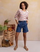 Woman wearing a colorful striped shirt and denim shorts standing in front of a wooden crate with fruits and plants.