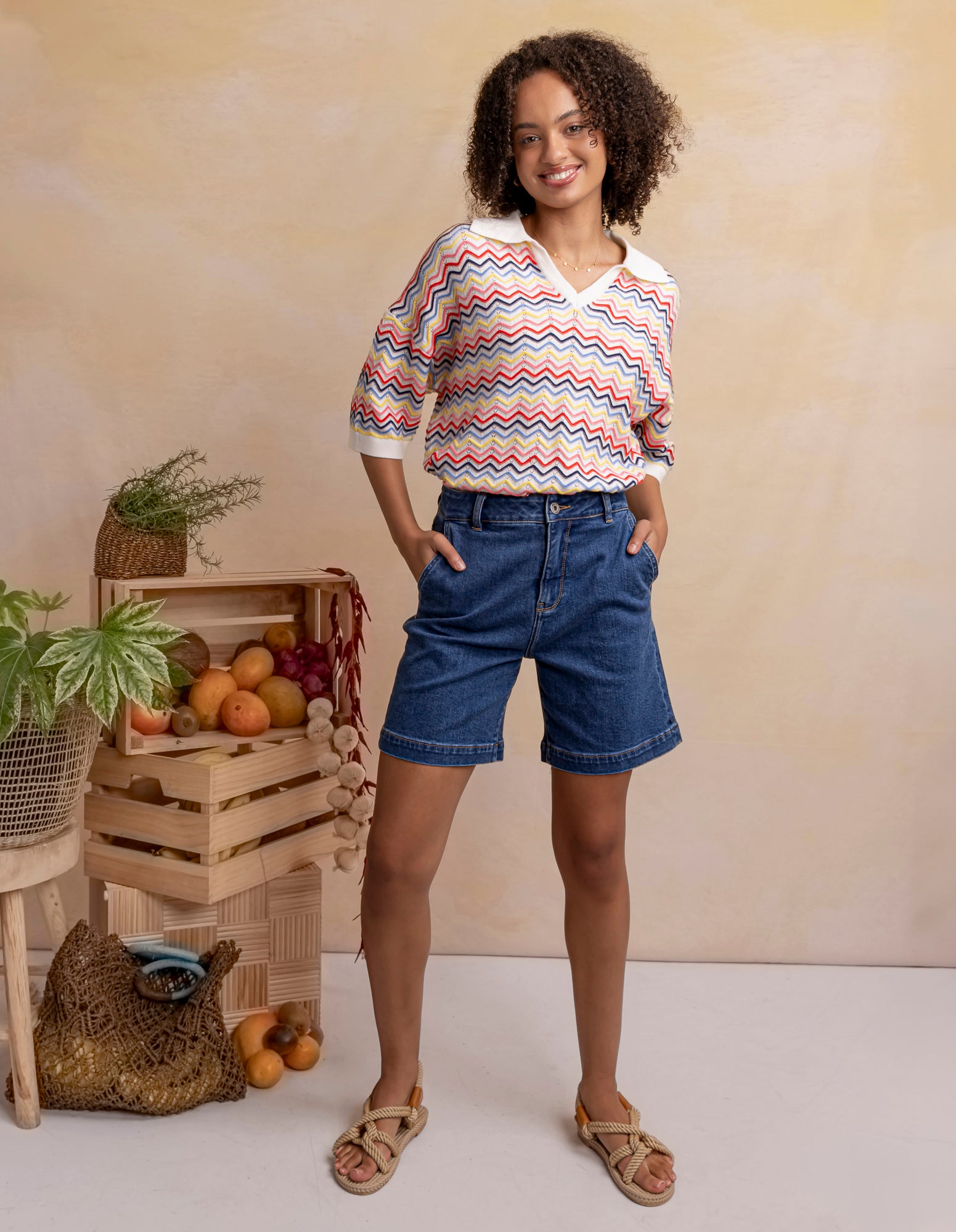 Woman wearing a colorful striped shirt and denim shorts standing in front of a wooden crate with fruits and plants.
