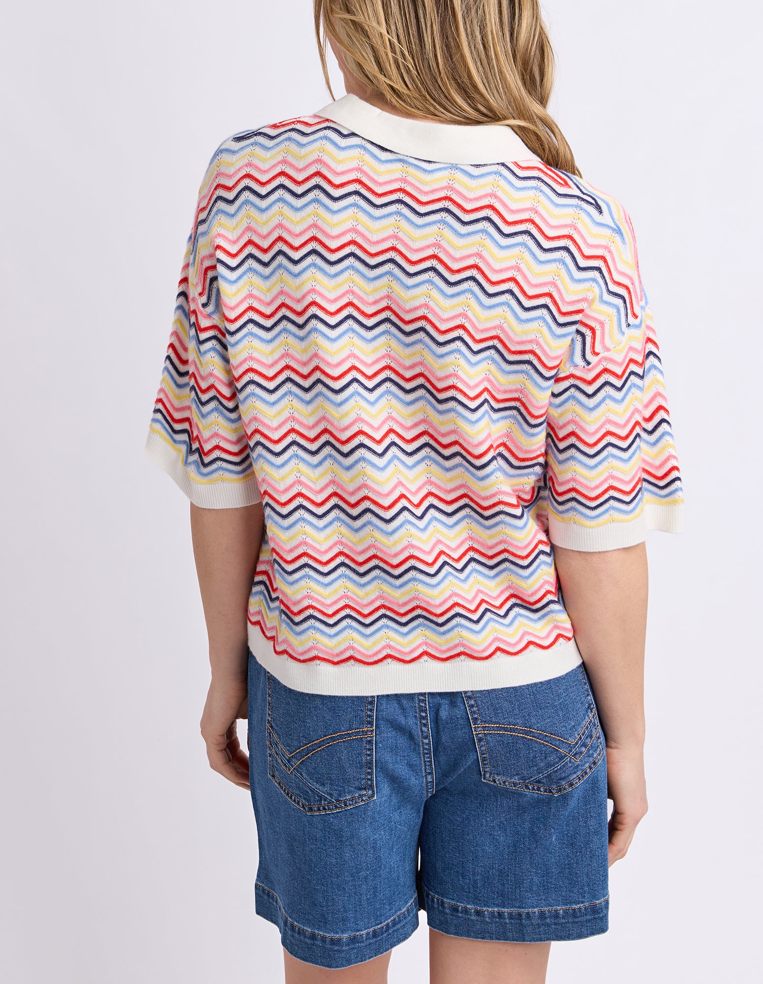 A child model shown from the back wearing a short-sleeve knit polo shirt with a multicolor chevron pattern and blue denim shorts against a light gray studio background.