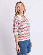A woman wearing a short-sleeve knit top with a colorful chevron pattern in red, blue, yellow, and white, paired with blue denim shorts, photographed against a light gray studio background.