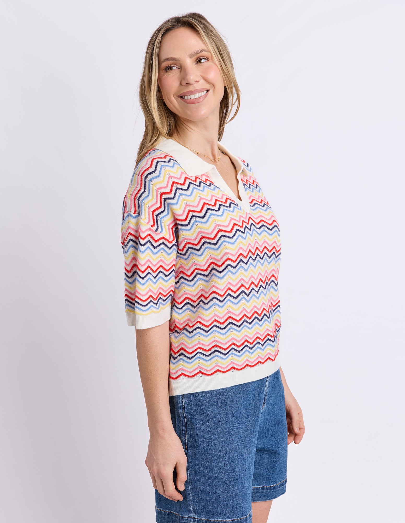 A woman wearing a short-sleeve knit top with a colorful chevron pattern in red, blue, yellow, and white, paired with blue denim shorts, photographed against a light gray studio background.