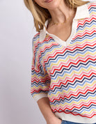 A close-up of a woman wearing a colorful zigzag knit polo sweater with a cream collar and cuffs, paired with a delicate gold necklace.