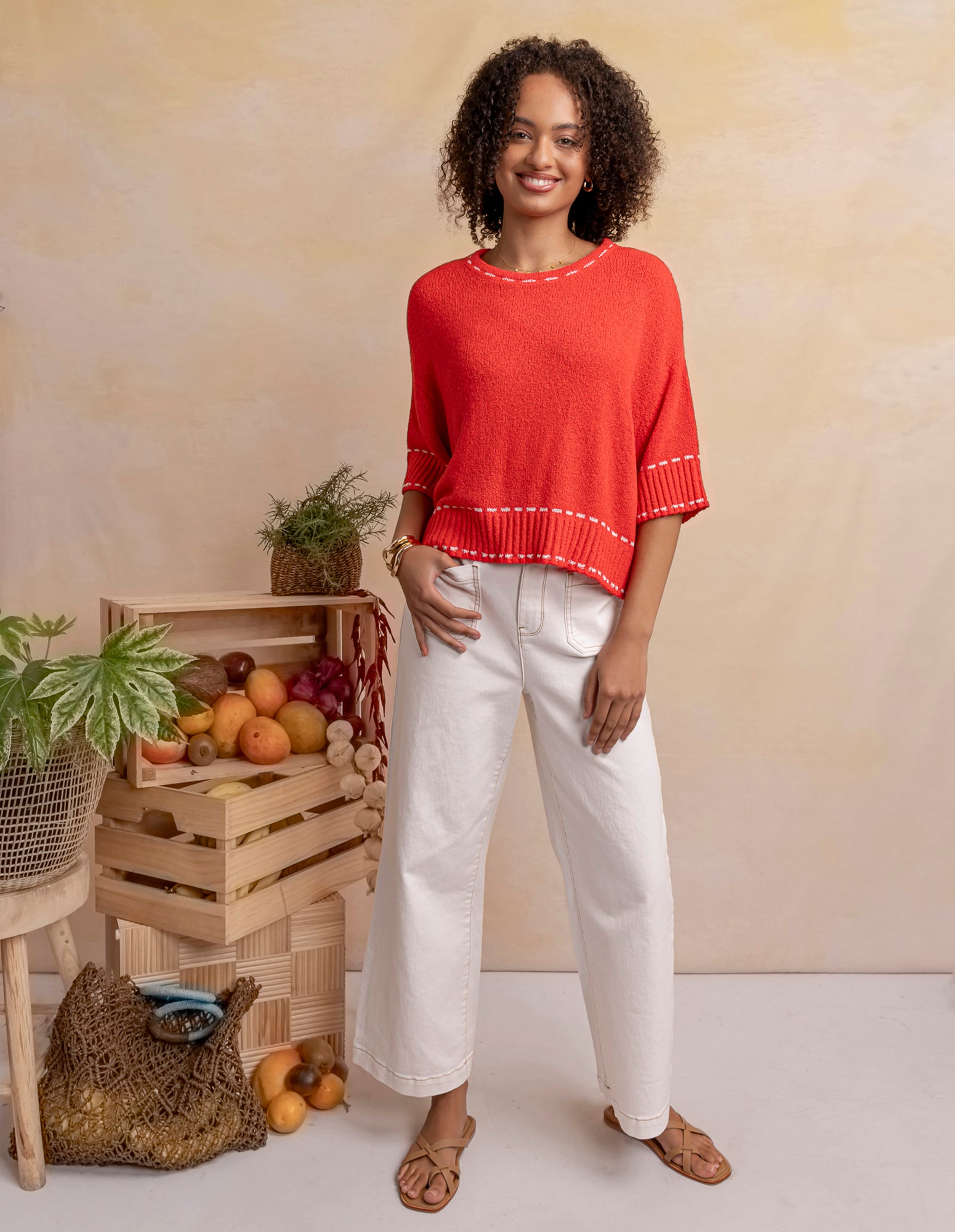 Woman in a red sweater and white pants standing next to wooden crates with fruits and plants.