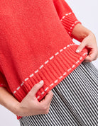 Red sweater with white trim held by a person wearing striped pants on a light gray background