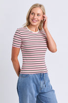 A woman modeling a short-sleeve crewneck t-shirt with horizontal pink, black, and white stripes, paired with light blue jeans, against a light gray studio background.