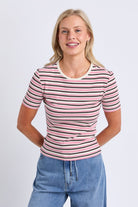 A woman modeling a short-sleeve ribbed knit top with horizontal pink, black, and white stripes, paired with blue jeans, against a light gray studio background.