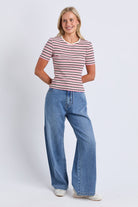 A woman modeling a short-sleeve crewneck t-shirt with thin red and navy horizontal stripes, paired with wide-leg blue jeans and white sneakers, against a light gray studio background.