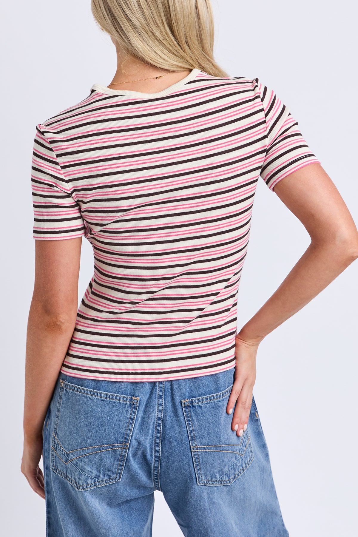 A woman shown from the back wearing a short-sleeve striped t-shirt with pink, black, and white horizontal stripes, paired with blue jeans, against a light gray studio background.