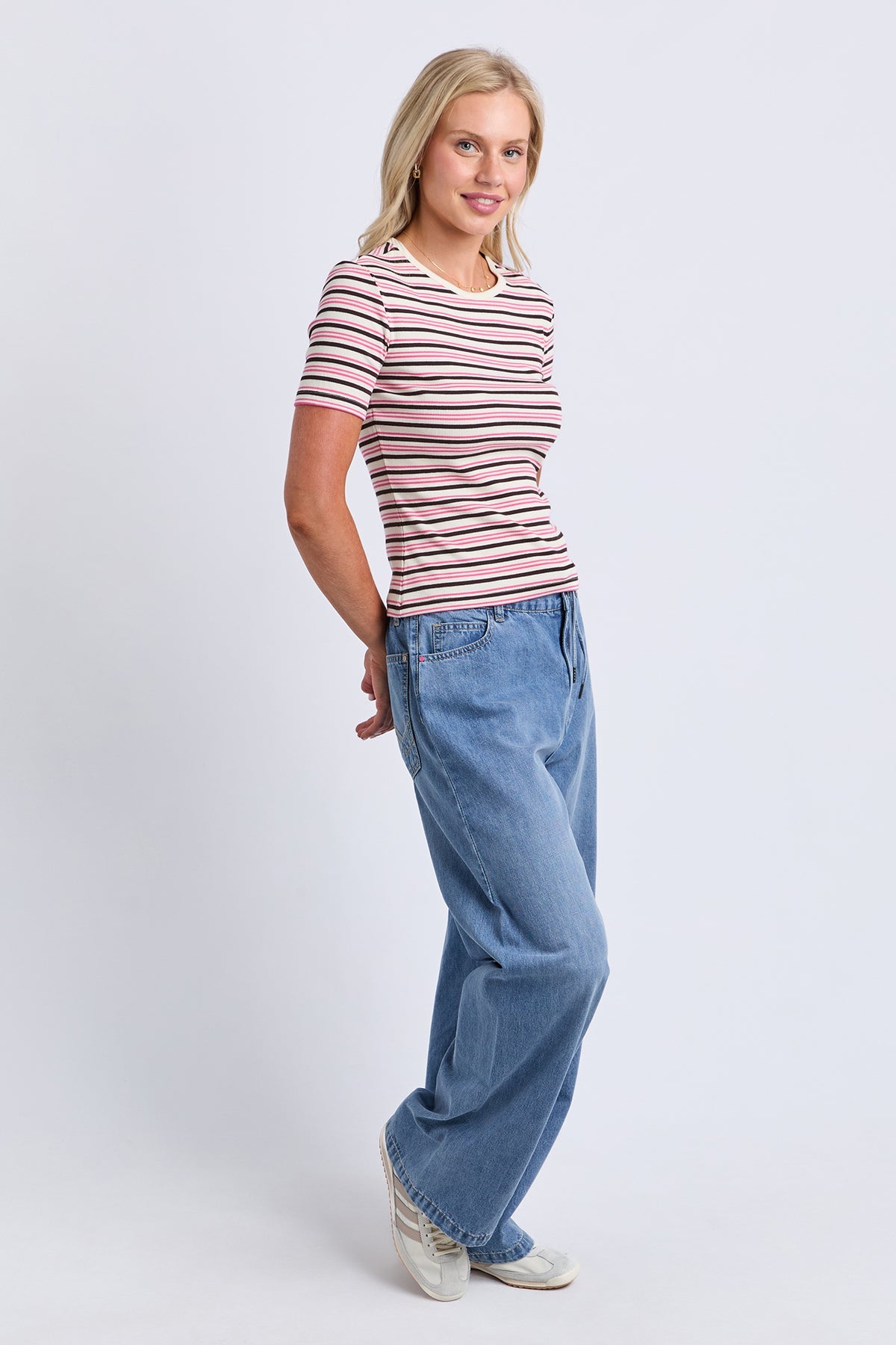 A woman modeling a short-sleeve crewneck t-shirt with thin pink and navy horizontal stripes, paired with wide-leg blue jeans and white sneakers, against a light gray studio background.