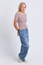 A woman modeling a short-sleeve crewneck t-shirt with thin pink and navy horizontal stripes, paired with wide-leg blue jeans and white sneakers, against a light gray studio background.