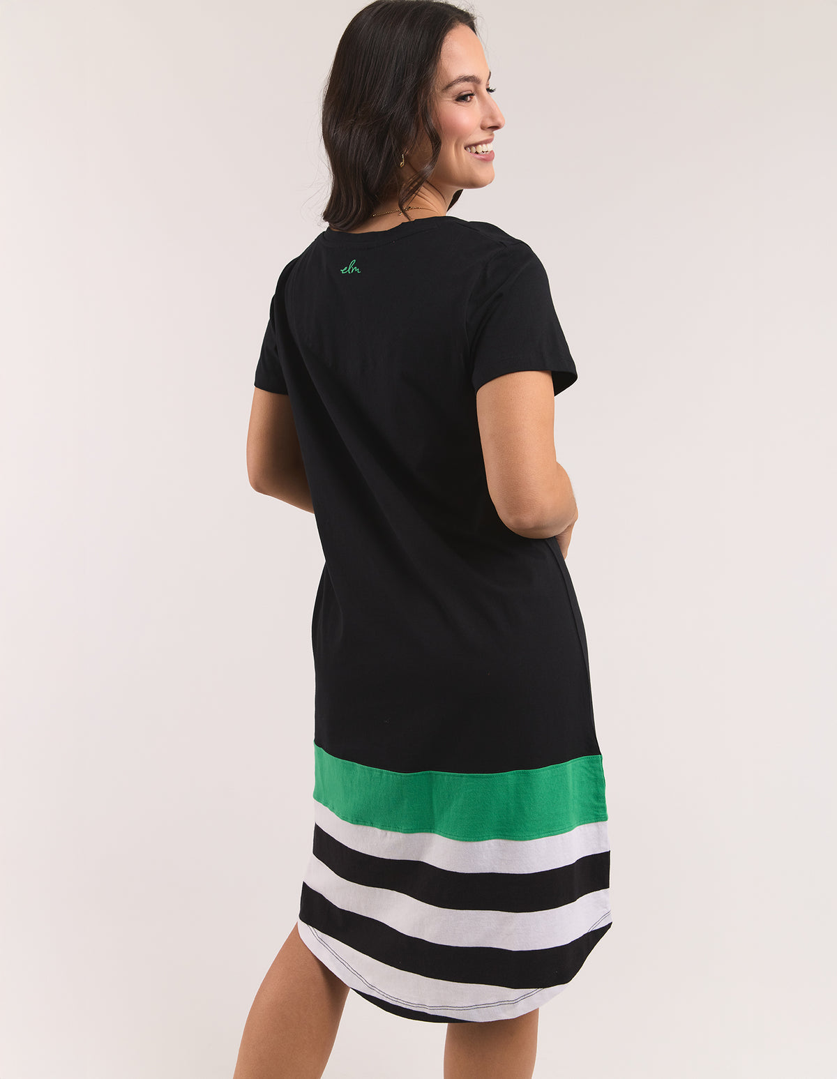 A woman modeling a black short-sleeve dress with a green band and black, white, and green horizontal stripes at the hem, shown from a three-quarters back view against a light beige studio background.