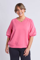 A woman wearing a bright pink V-neck sweatshirt with elbow-length balloon sleeves and elastic cuffs, paired with dark leggings, against a light gray studio background.