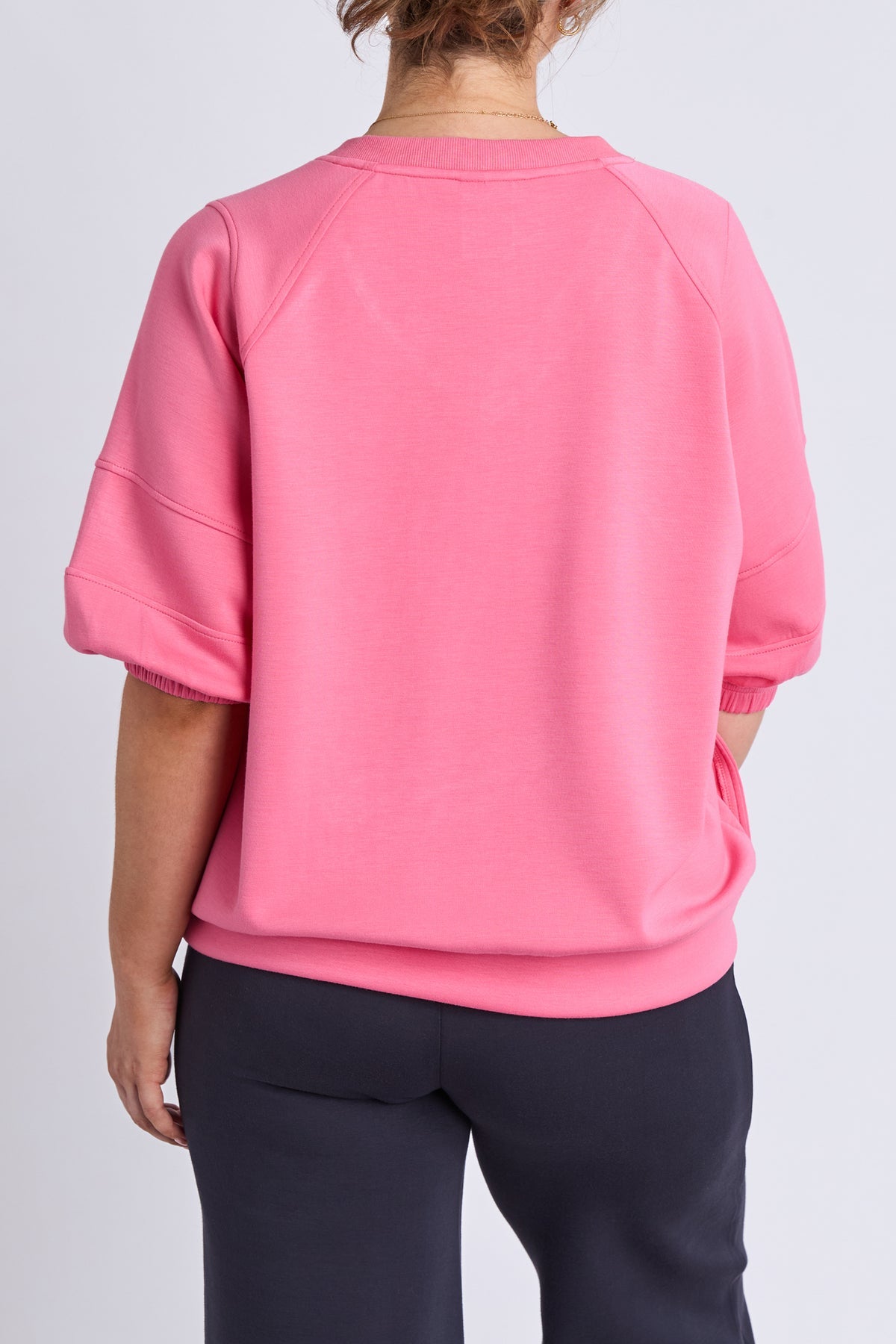 A person shown from the back wearing a bright pink short-sleeve sweatshirt and dark navy pants against a light gray studio background.