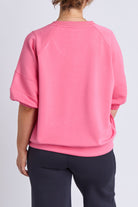 A person shown from the back wearing a bright pink short-sleeve sweatshirt and dark navy pants against a light gray studio background.