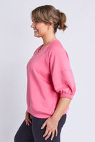 A woman wearing a pink short-sleeve sweatshirt with elasticized cuffs and a V-neck, paired with dark pants, shown in profile against a light gray studio background.