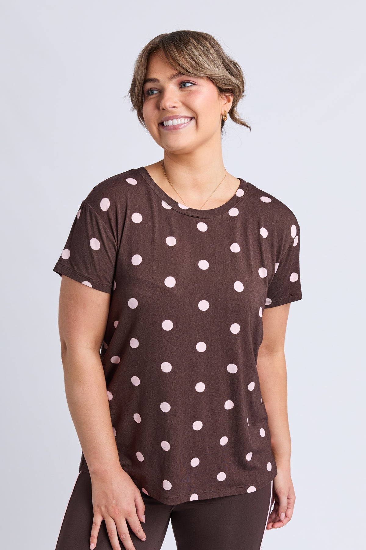 A woman wearing a brown short-sleeve t-shirt with a white polka dot pattern, paired with dark brown leggings, standing against a light gray studio background.