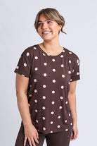 A woman wearing a brown short-sleeve t-shirt with a white polka dot pattern, paired with dark brown leggings, standing against a light gray studio background.