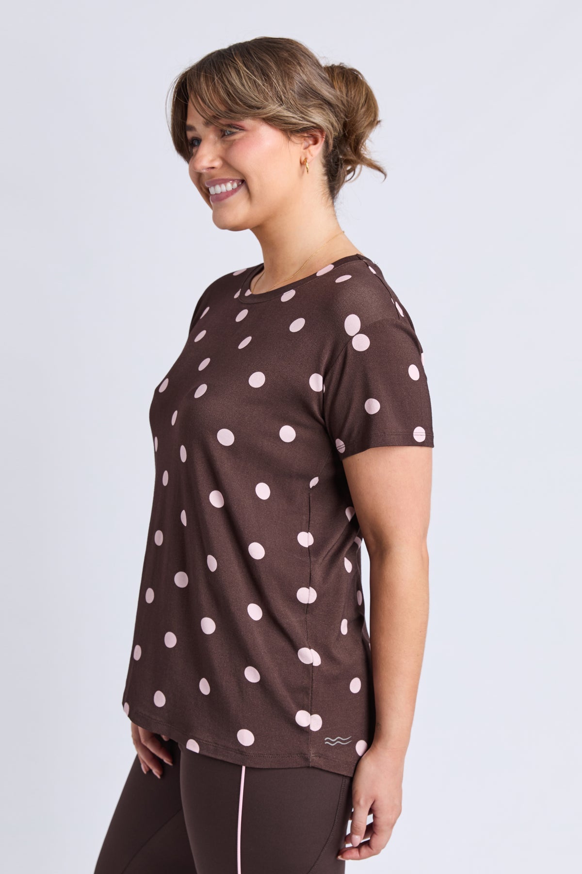 A woman wearing a brown short-sleeve athletic top with a white polka dot pattern, shown from a side profile against a light gray studio background.