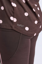 Close-up of a person wearing dark brown leggings and a matching dark brown top with large light pink polka dots and a small wavy line logo near the hem.