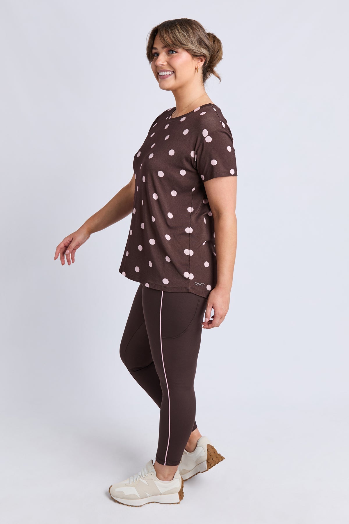 A woman modeling a brown short-sleeve t-shirt with white polka dots and matching brown leggings with a white side stripe, paired with white sneakers, against a light gray studio background.