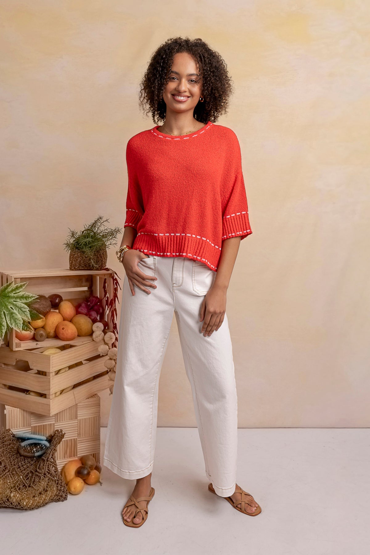 A woman stands in a studio wearing a bright coral-red knit poncho-style top with white contrast stitching and white wide-leg pants, paired with tan sandals. To her left is a rustic crate filled with fruit and small plants against a warm beige textured backdrop.