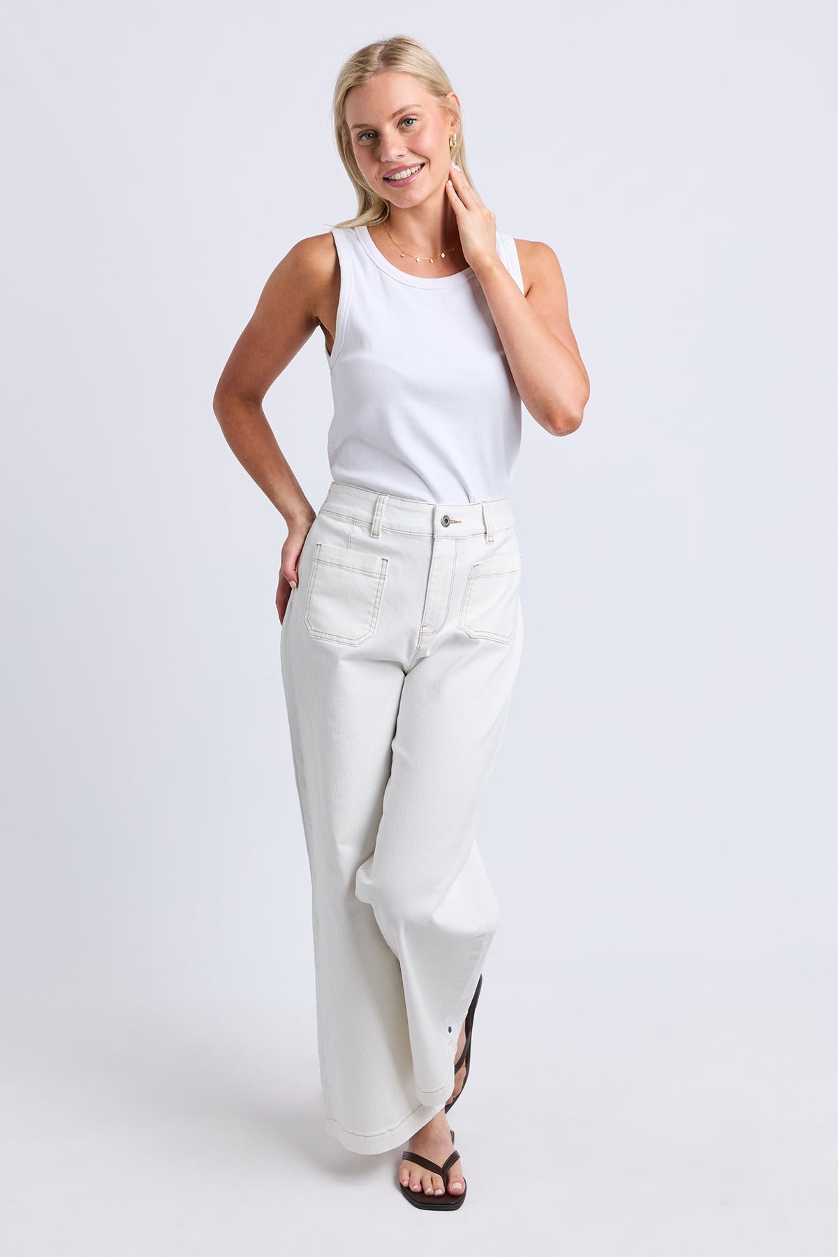 A woman modeling a white sleeveless tank top and high-waisted, wide-leg white jeans against a light gray studio background.