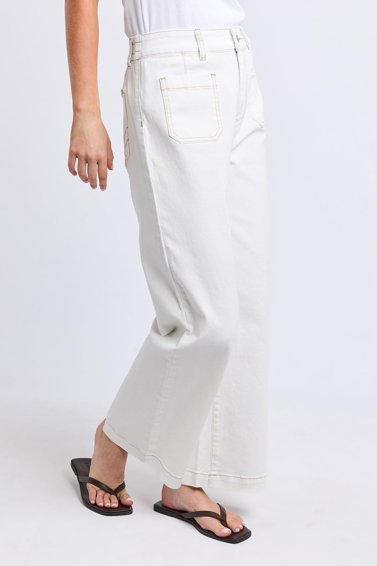 A person wearing off-white cropped wide-leg jeans with a back pocket, paired with black flip-flops, shown from the waist down against a light gray studio background.
