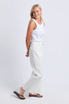 A woman modeling a pair of high-waisted, wide-leg white jeans with a white tank top and black flip-flops, standing against a light gray studio background.