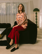 Woman sitting on a dark armchair wearing a patterned sweater and red pants in a living room.