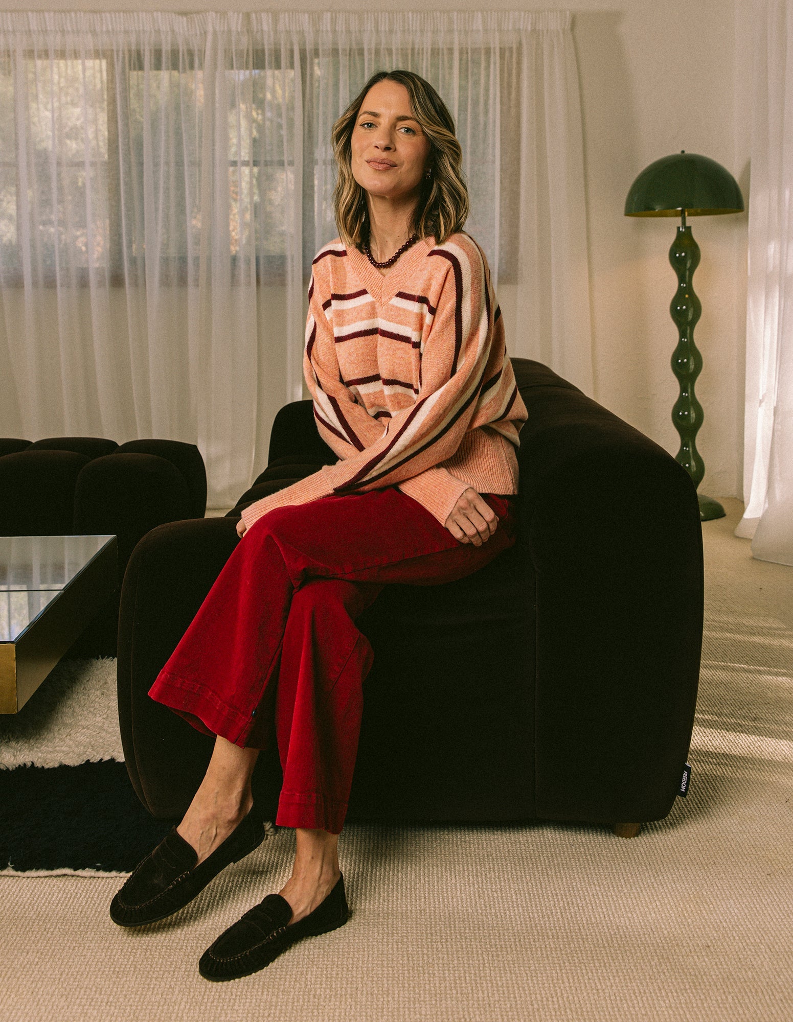 Woman sitting on a dark armchair wearing a patterned sweater and red pants in a living room.