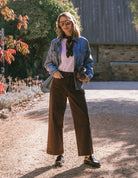 Woman standing outdoors wearing a denim jacket, white shirt, brown pants, and black shoes.