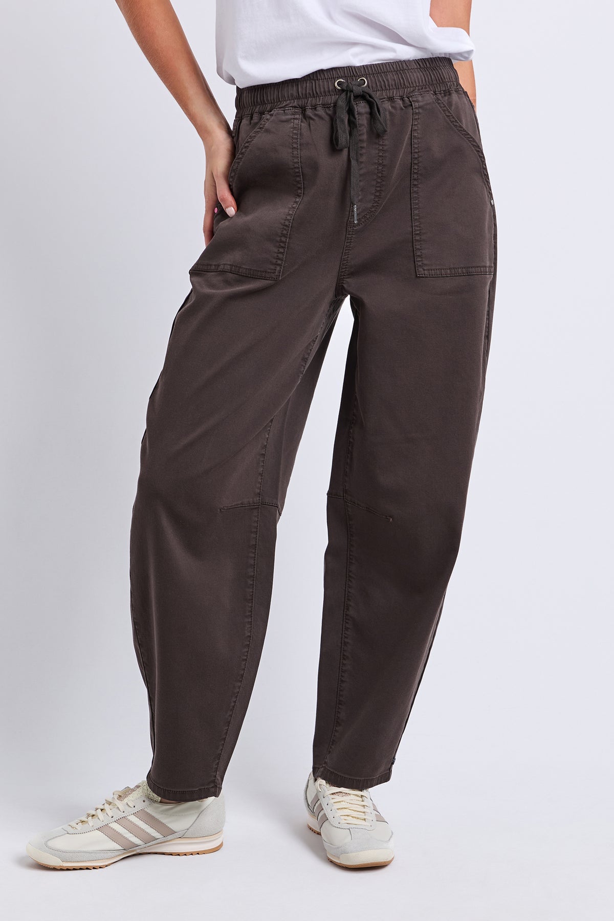 A person wearing dark brown drawstring pants with side pockets, paired with a white top and beige sneakers, shown from the waist down against a light gray studio background.