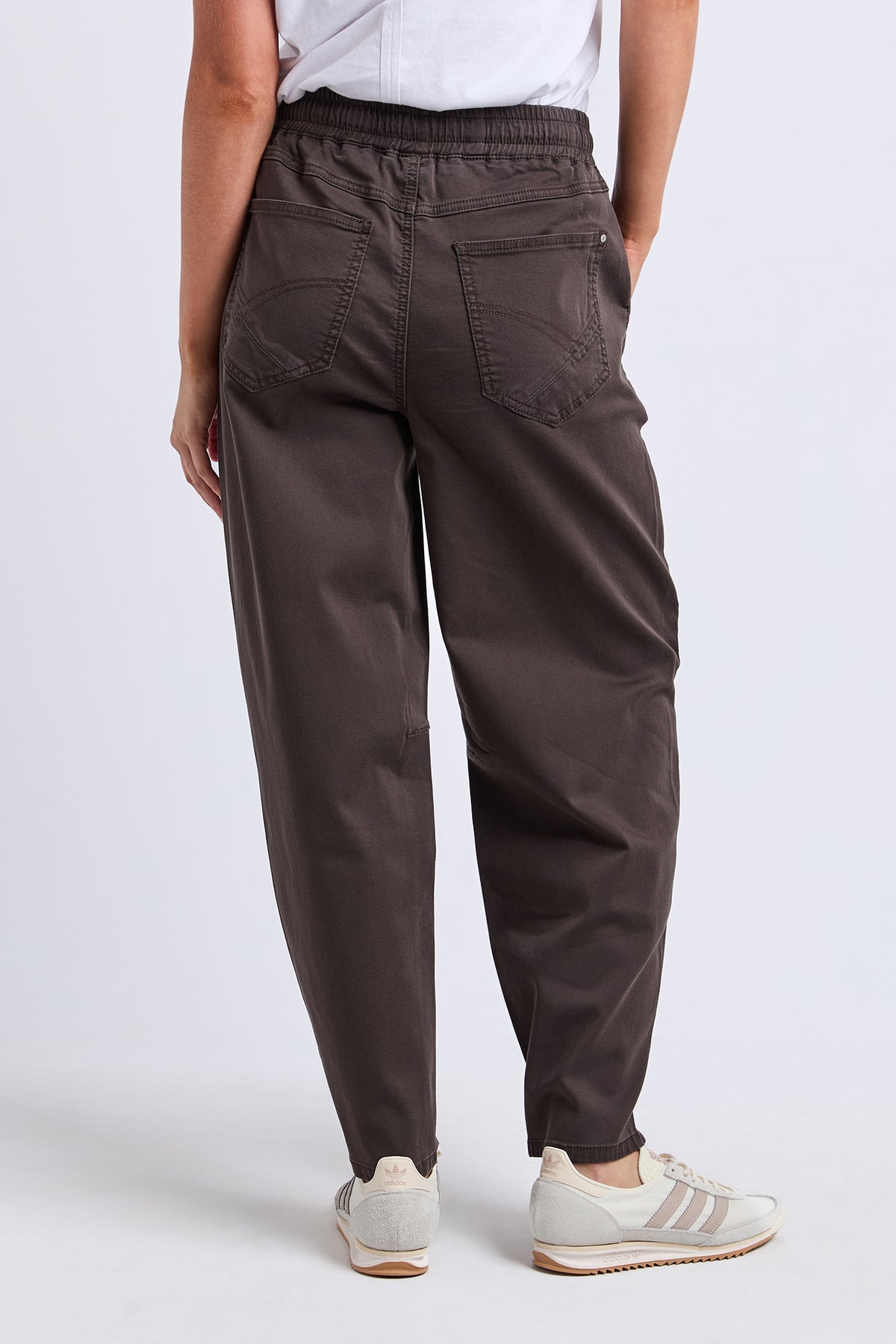 A person shown from the back wearing dark brown tapered pants with an elastic waistband, rear patch pockets, and light beige sneakers against a light gray studio background.