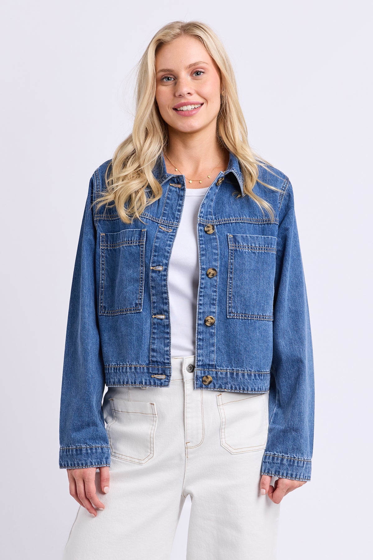 A blonde woman wearing a cropped blue denim jacket over a white top and white pants, standing against a light gray studio background.