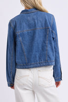 A person shown from the back wearing a cropped blue denim jacket with white pants, photographed against a clean light gray studio background.