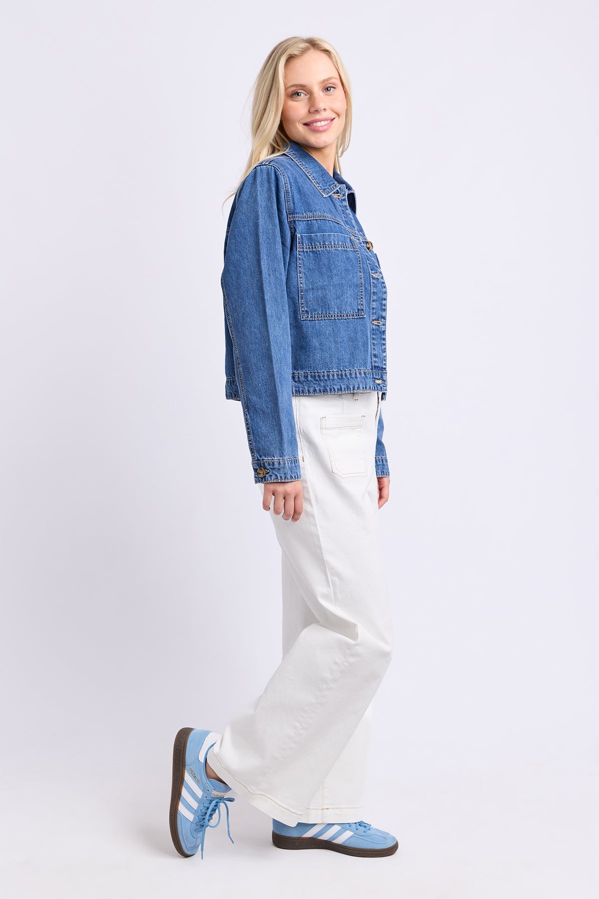 A woman modeling a cropped blue denim jacket, white wide-leg jeans, and light blue sneakers against a plain light gray studio background.
