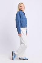 A woman modeling a cropped blue denim jacket, white wide-leg jeans, and light blue sneakers against a plain light gray studio background.
