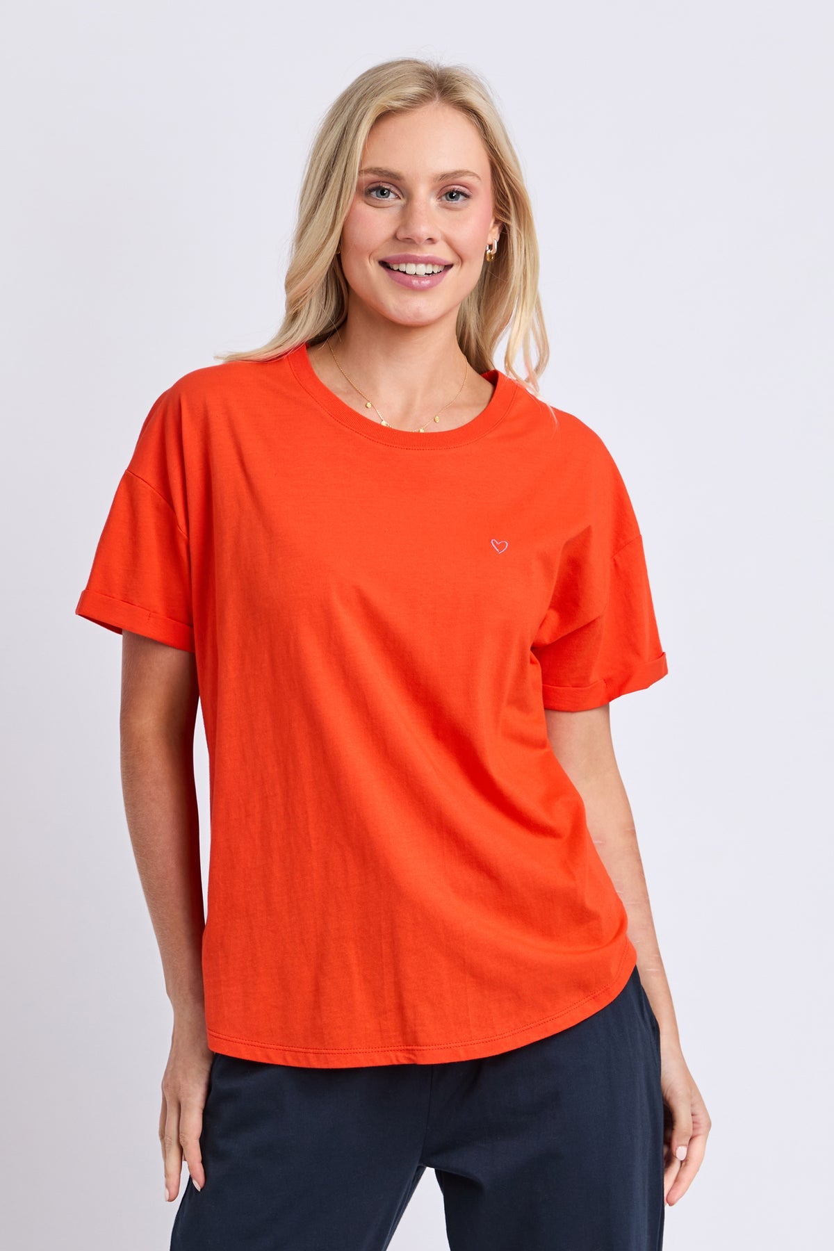 A woman modeling a bright orange short-sleeve crewneck t-shirt with a small red heart logo on the left chest, paired with navy pants, against a light gray studio background.