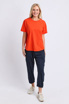 A woman modeling a bright orange short-sleeve crewneck t-shirt and dark navy cropped pants with white sneakers, standing against a light gray studio background.