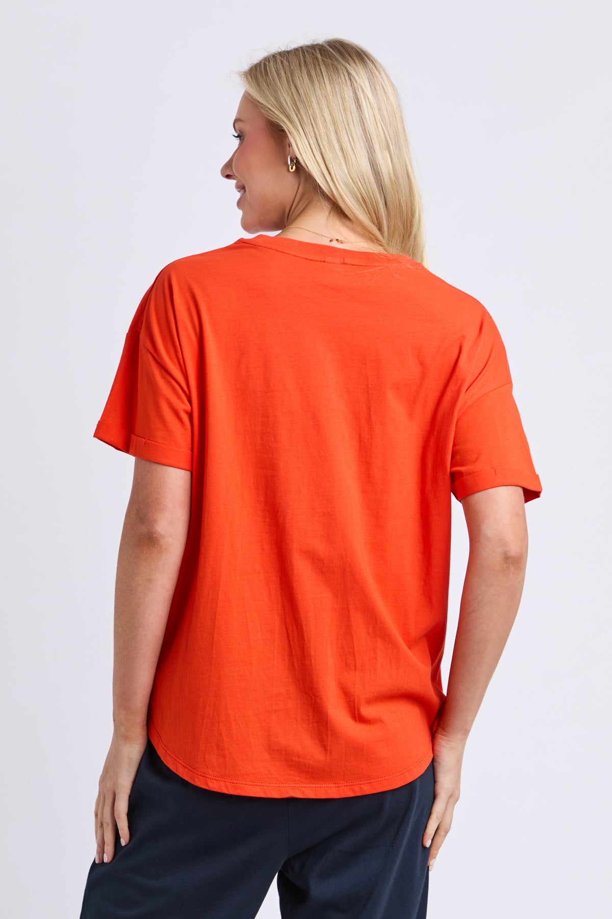 A woman shown from the back wearing a bright orange short-sleeve t-shirt and navy blue pants, photographed against a light gray studio background.