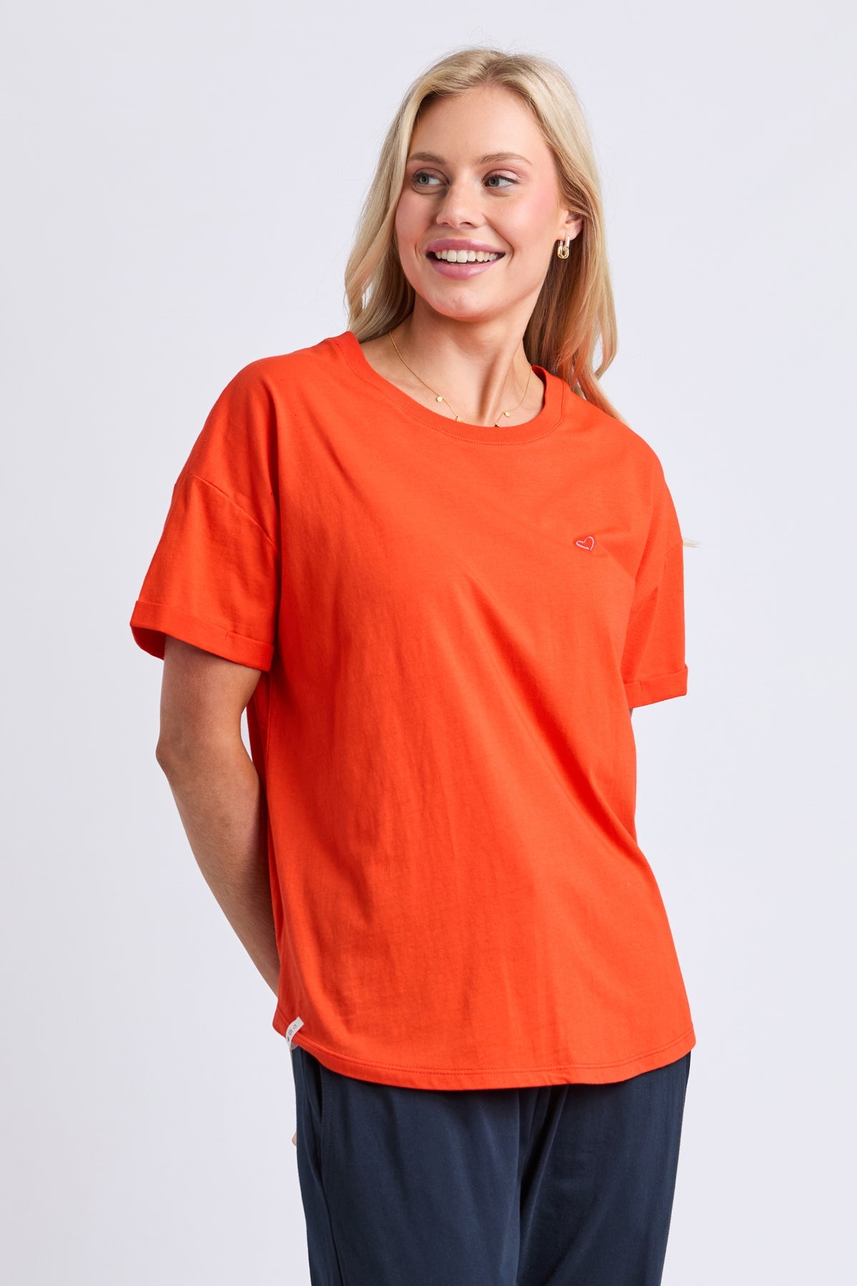 A smiling blonde woman wearing a bright orange short-sleeve crewneck t-shirt with a small chest pocket and navy blue pants, photographed against a light gray studio background.