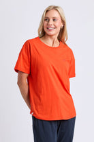 A smiling blonde woman wearing a bright orange short-sleeve crewneck t-shirt with a small chest pocket and navy blue pants, photographed against a light gray studio background.