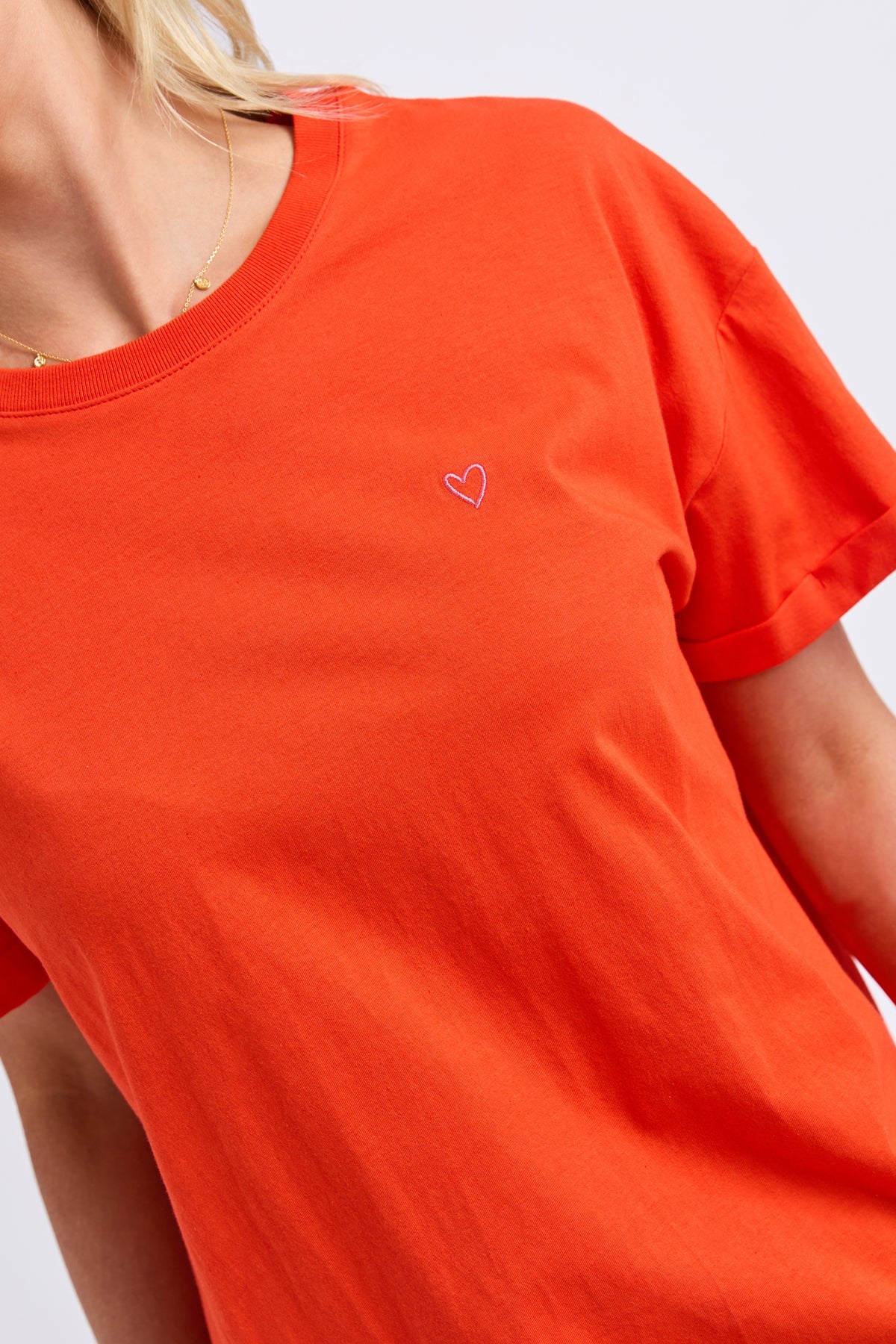 A close-up of a woman wearing a bright orange short-sleeve crewneck t-shirt with a small white heart outline embroidered on the left chest.