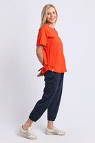 A woman modeling a bright orange short-sleeve t-shirt and navy blue jogger pants with white sneakers, standing against a light gray studio background.